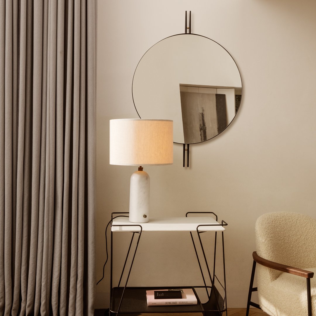 Modern round mirror with a black frame, neutral beige lamp, sleek white table, and soft gray curtains. Elegant and minimalist design.
