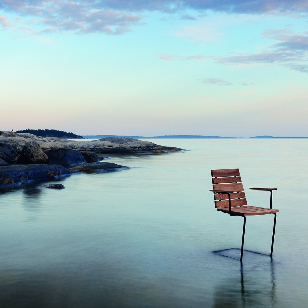 Brown slatted outdoor chair with a sleek metal frame, set against a serene blue water and pastel sky background.