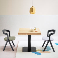 Modern dining set with a wooden table, black legs, green and gray chairs, and a pastel-patterned rug. Gold pendant light above.