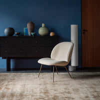 "Modern beige chair with gold legs on a soft, neutral rug, against a deep blue wall and dark wooden furniture."