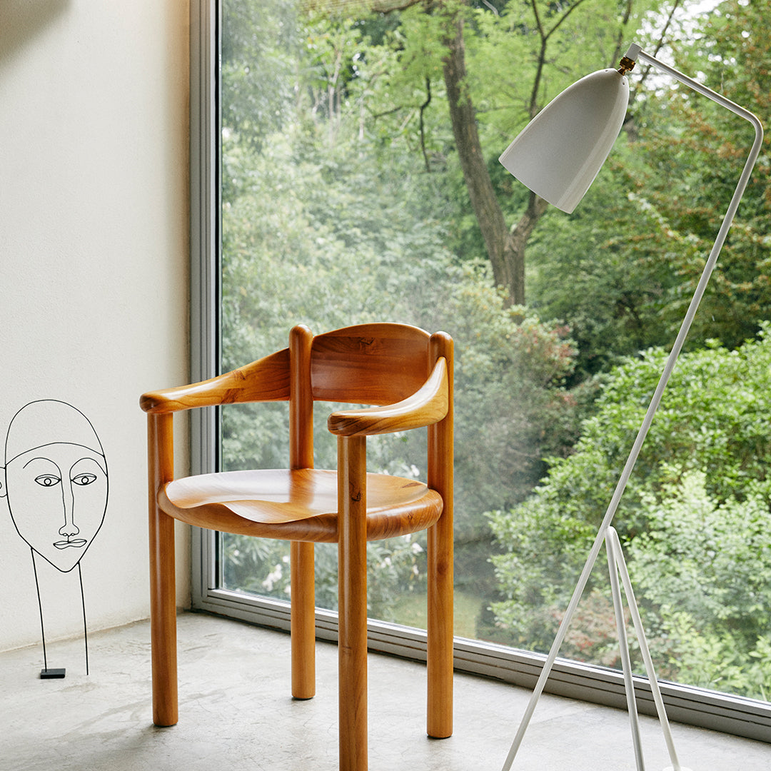 Natural wood chair with a smooth finish, placed near a large window with greenery in the background. Minimalist design.