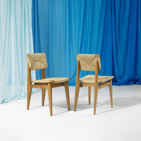 Pair of stylish mid-century chairs with a textured beige seat against a backdrop of gradient blue drapes.