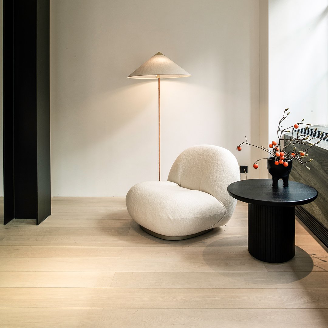 Plush cream-colored chair with a smooth, organic shape, paired with a sleek black side table and warm wooden flooring.