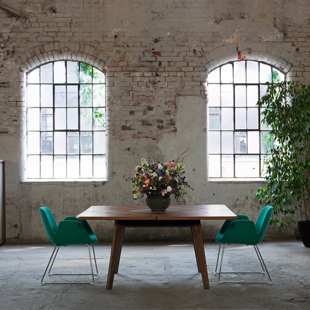 Modern wooden table with teal chairs, vibrant floral arrangement, and rustic brick walls in a bright space.