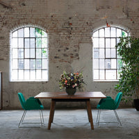 Modern wooden table with teal chairs, vibrant floral arrangement, and rustic brick walls in a bright space.