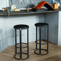 "Black metal bar stools with round seats and a minimalist design, set against a light wooden floor and textured background."
