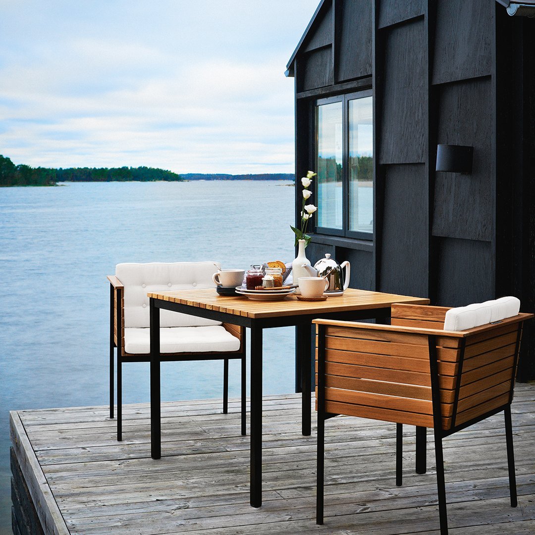 Elegant wooden dining table with black metal accents and creamy white cushions, set against a serene lake backdrop.