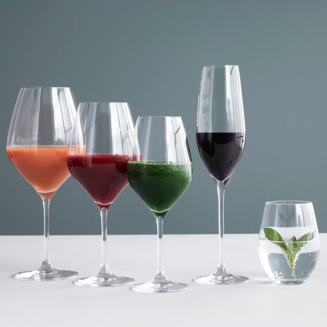 Set of five elegant wine glasses with vibrant juices: peach, berry, green, red, and clear water with mint leaf.