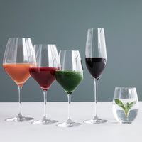 Set of five elegant wine glasses with vibrant juices: peach, berry, green, red, and clear water with mint leaf.