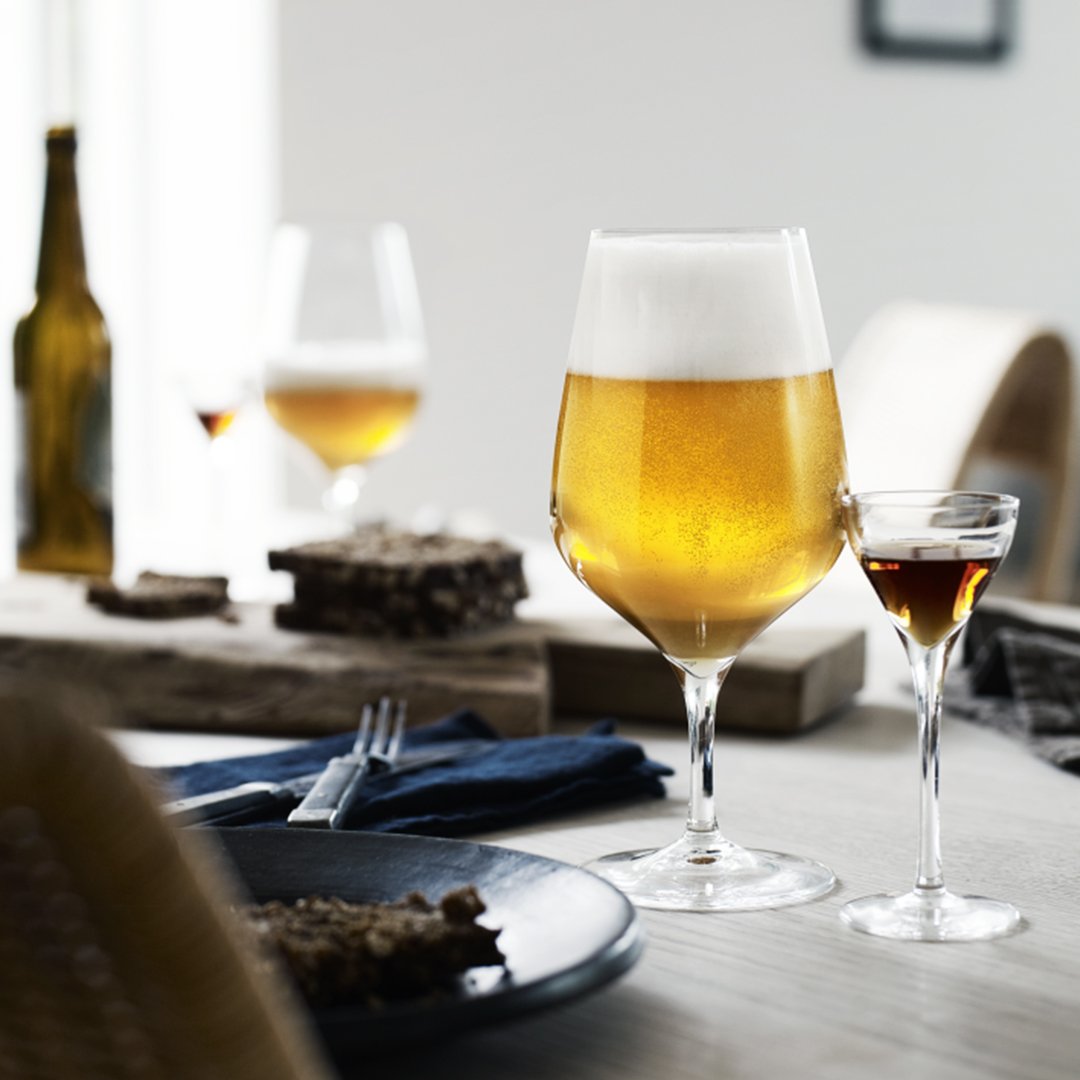 Golden beer in a large glass with a frothy top, accompanied by a smaller glass, set on a neutral-toned table.