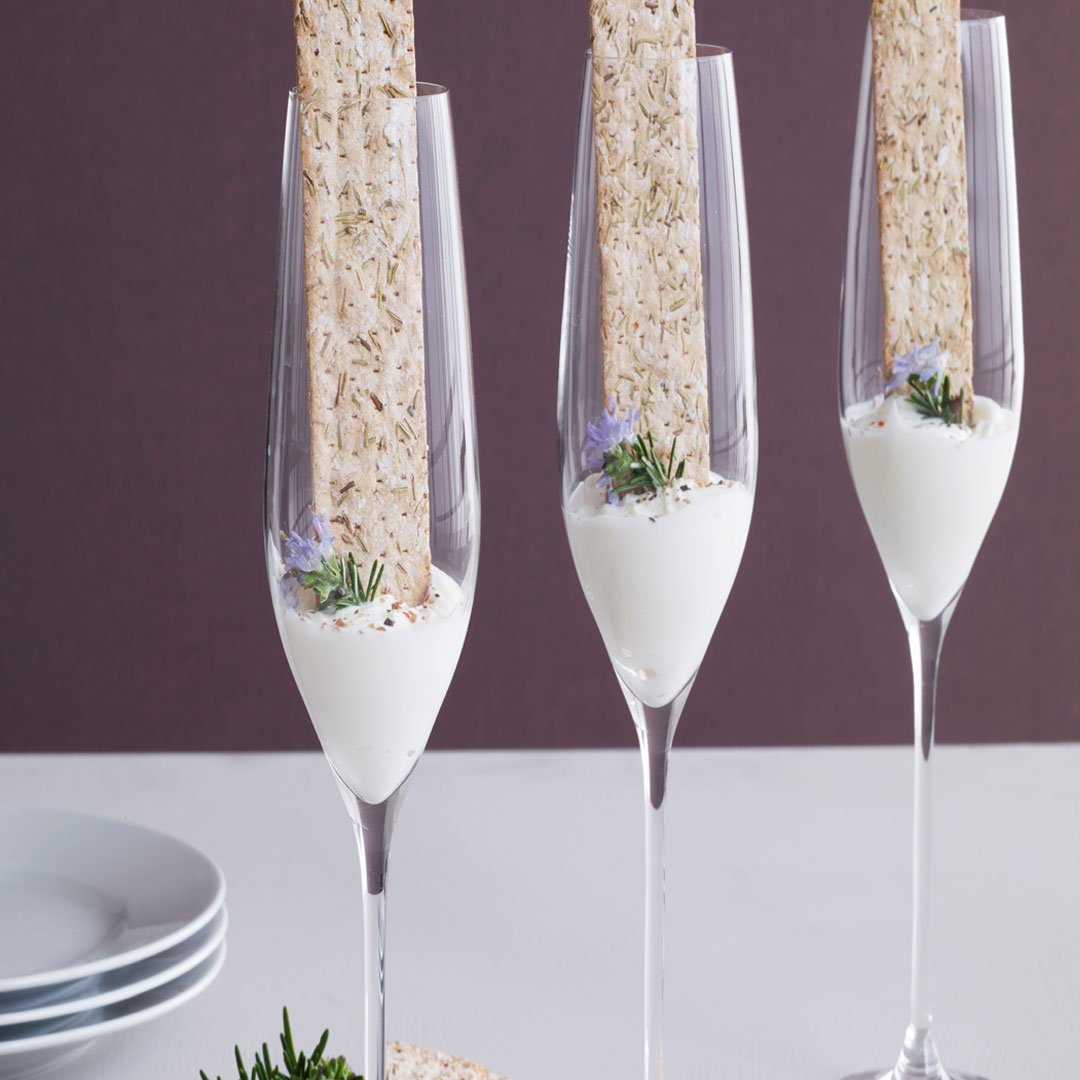 Elegant clear champagne flutes with white cream, topped with a beige cracker and sprigs of greenery on a neutral background.