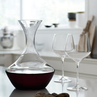 Elegant glass decanter with a sleek design, featuring a clear body and rich dark red wine at the base. Two clear wine glasses.