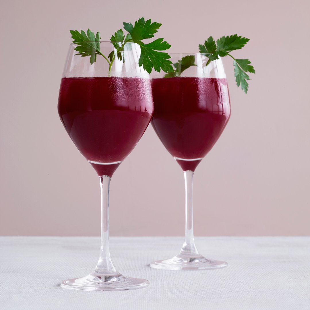 Two elegant glasses of deep burgundy drink garnished with fresh green parsley against a soft pink background.