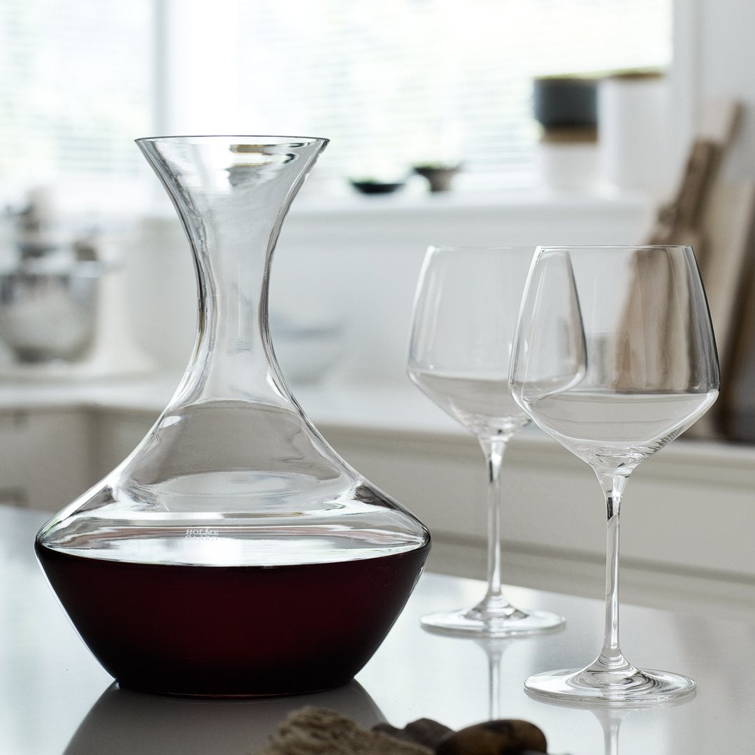 Elegant glass wine decanter with a curved shape, featuring a clear upper section and a rich red base, complemented by two clear glasses.