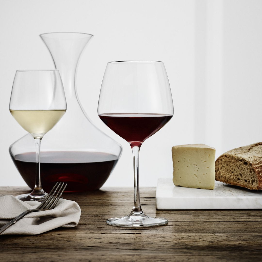 "Elegant wine glasses, one clear and one deep red, paired with a glass decanter and rustic bread on a wooden table."