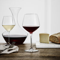 "Elegant wine glasses, one clear and one deep red, paired with a glass decanter and rustic bread on a wooden table."