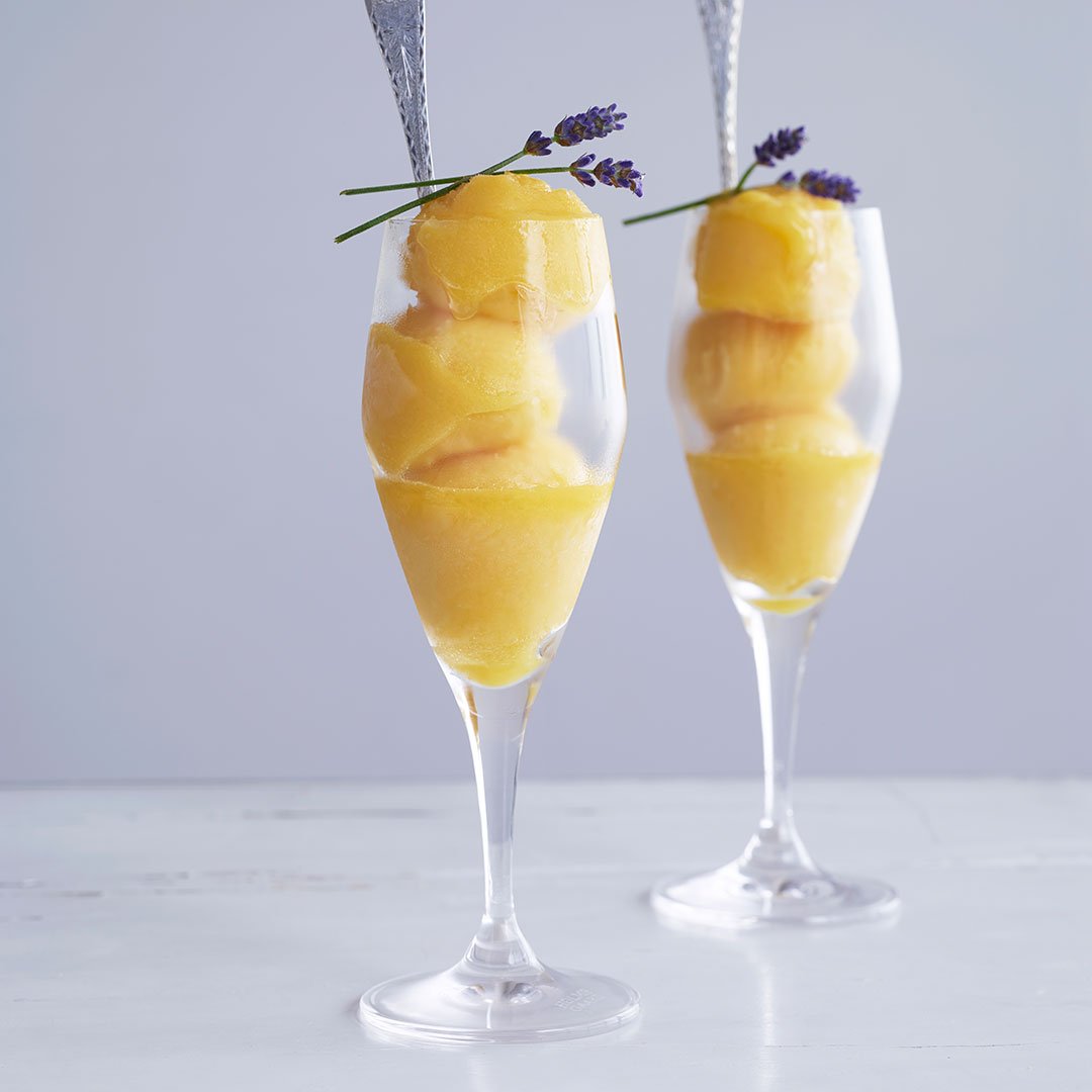 Two clear glasses filled with yellow sorbet, garnished with lavender sprigs, set against a light gray background.