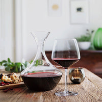 Elegant glass decanter and wine glass featuring deep red wine against a warm wooden table and soft green accents.