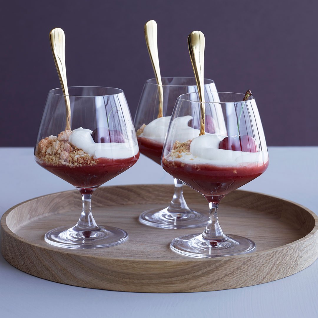 Elegant dessert in clear glasses with vibrant red fruit, creamy white topping, and golden spoons on a wooden tray.