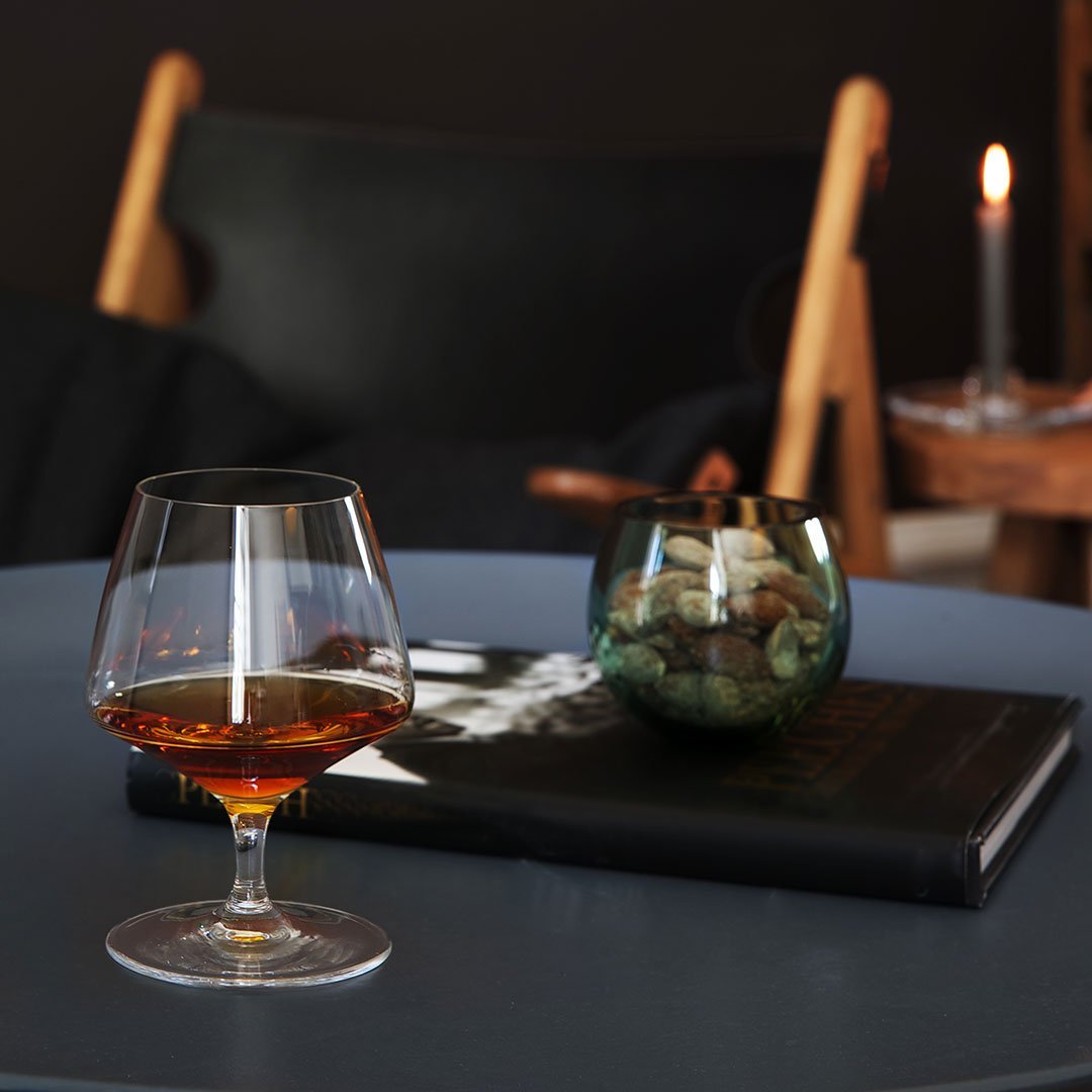 "Elegant glass with amber liquid, green glass bowl, and dark textured book on a round blue table."