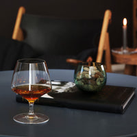 "Elegant glass with amber liquid, green glass bowl, and dark textured book on a round blue table."