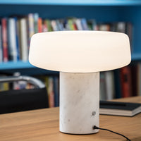Modern white lamp with a smooth marble base and a round, glossy shade, set against a colorful bookshelf backdrop.