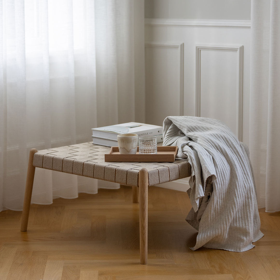 Light beige woven table with natural wood legs, accompanied by a striped gray throw and white decor.