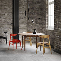 Modern round wooden table with natural finish, paired with red and light wood chairs, against a textured brick background.