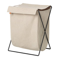 Beige fabric laundry basket with a black metal frame and a brown leather strap accent. Minimalist design.
