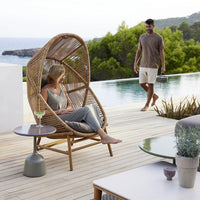Natural rattan chair with beige cushions, gray side table, and green patio decor amidst a serene outdoor setting.