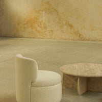 Cream-colored textured chair with a smooth beige stone coffee table against a muted warm-toned wall.