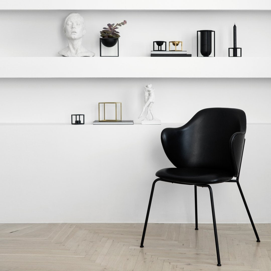Black chair with a smooth, modern design, set against a minimalist white wall and light wooden floor.