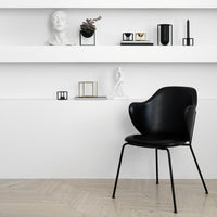 Black chair with a smooth, modern design, set against a minimalist white wall and light wooden floor.