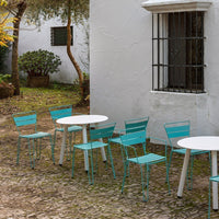 "Set of turquoise metal chairs and a round white table against a textured stone patio and white wall."