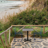 Outdoor dining set featuring a gray table and blue wire chairs against a natural stone patio backdrop.