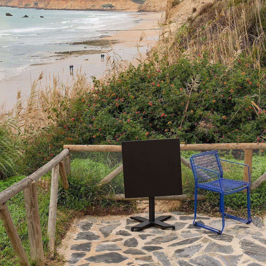 Black square table with a modern design next to a vibrant blue wire chair on a stone patio with a scenic beach backdrop.