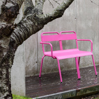 Bright pink metal bench with a sleek, modern design, set against a neutral concrete background and tree branches.