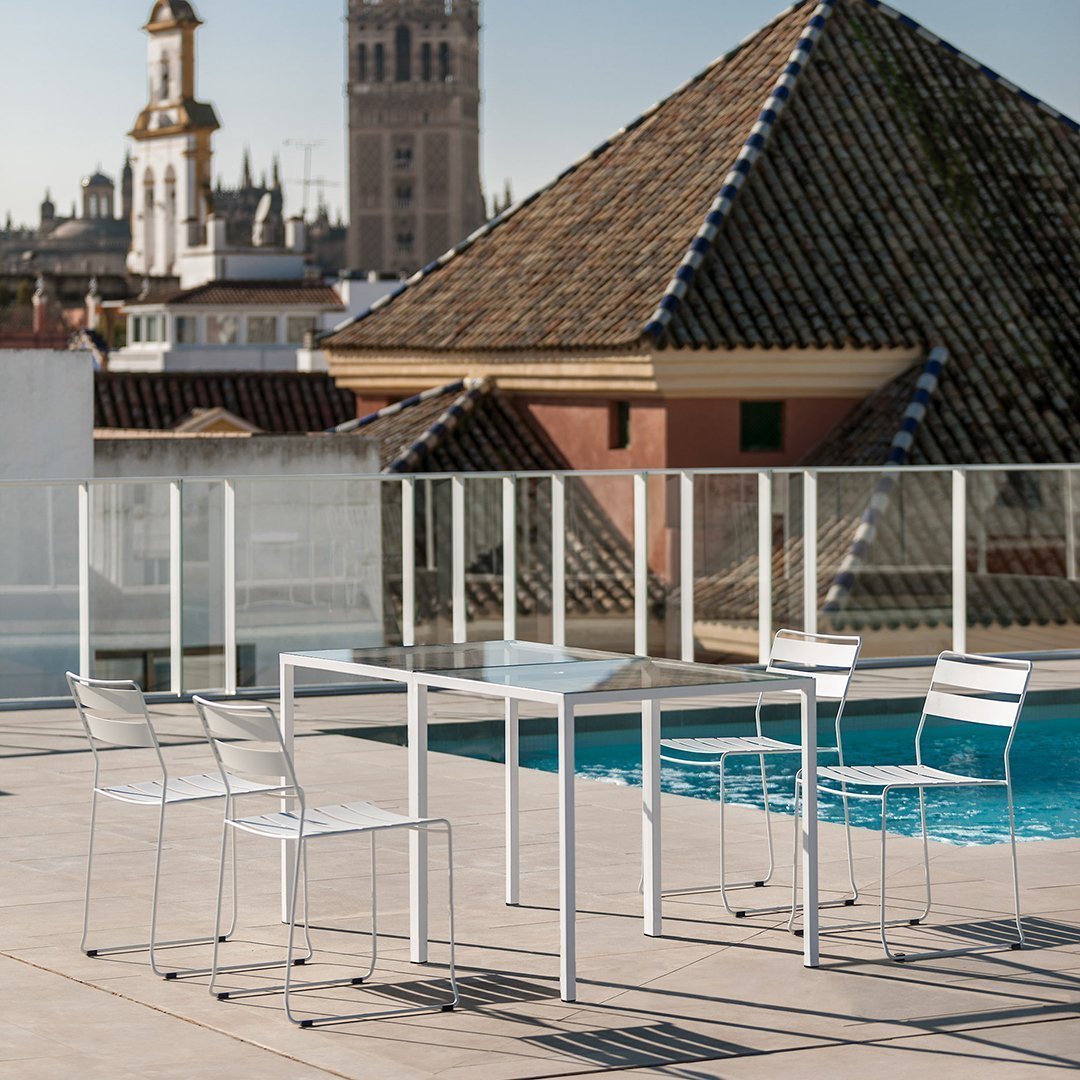 White outdoor dining table with sleek lines, complemented by four matching transparent chairs against a blue pool backdrop.