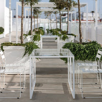 "White glass-top table with minimalist design, surrounded by greenery and sleek white chairs in a modern outdoor setting."