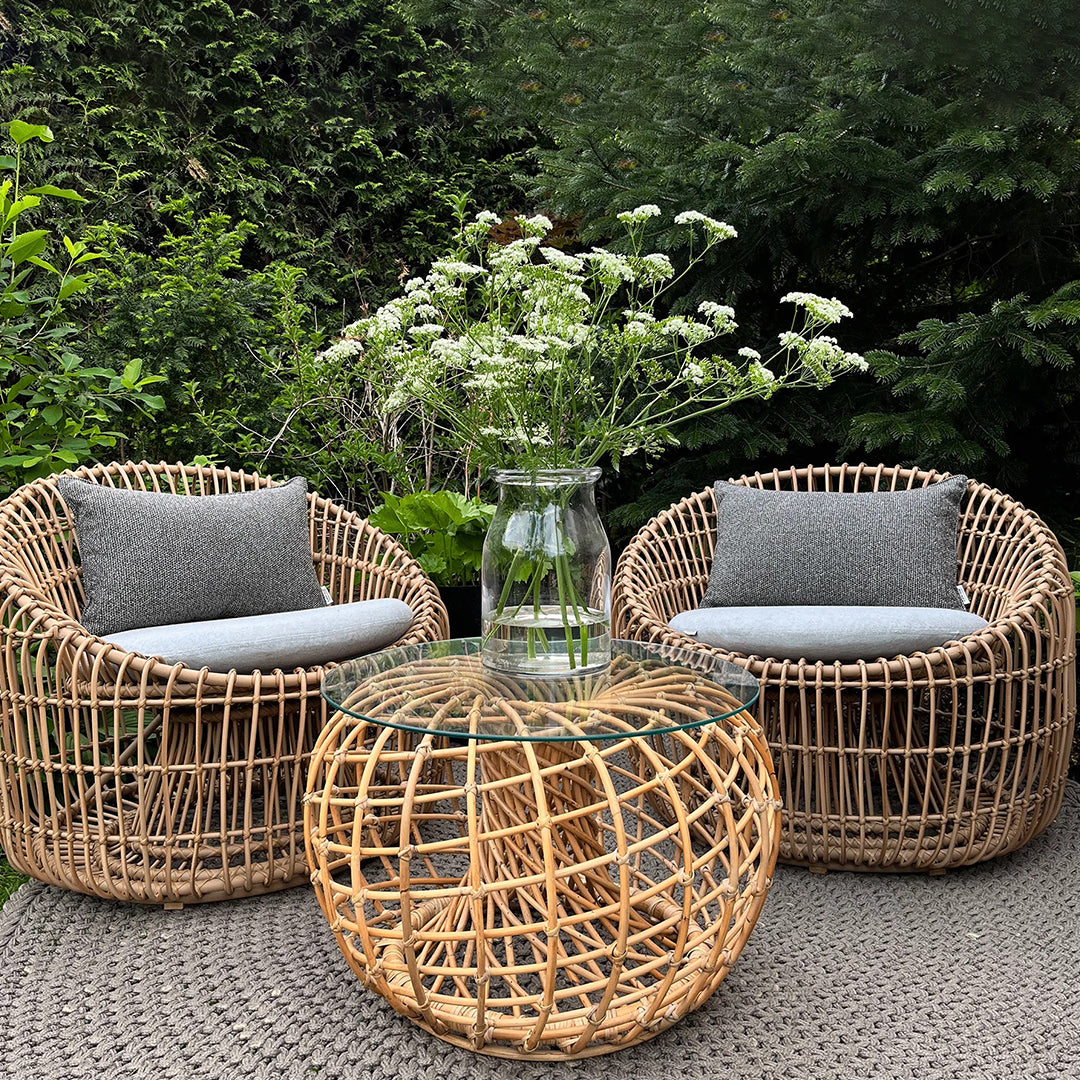 Natural wicker chairs with gray cushions and a rounded glass-top table, set on a textured gray outdoor rug.
