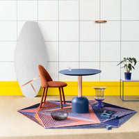 Colorful geometric rug in blue, orange, and purple, paired with a modern blue table and rust-colored chair against a white wall.