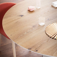 Round wooden table with a light oak finish, featuring a beige and gold accent plate, complemented by a coral chair.