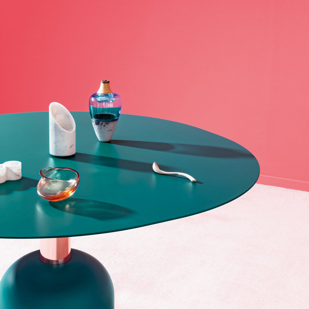 Teal round table with coral wall backdrop, showcasing decorative glass items in vibrant colors and unique patterns.