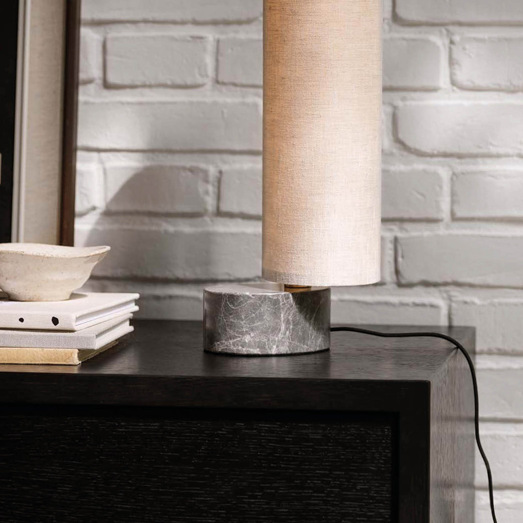 Modern table lamp with a textured beige shade and a sleek gray marble base, set against a white brick wall.