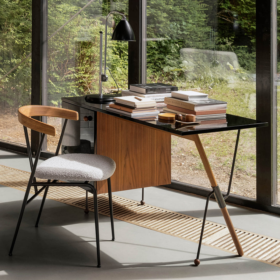 Modern desk with a sleek black surface, warm wooden accents, and a gray upholstered chair in a bright, natural setting.