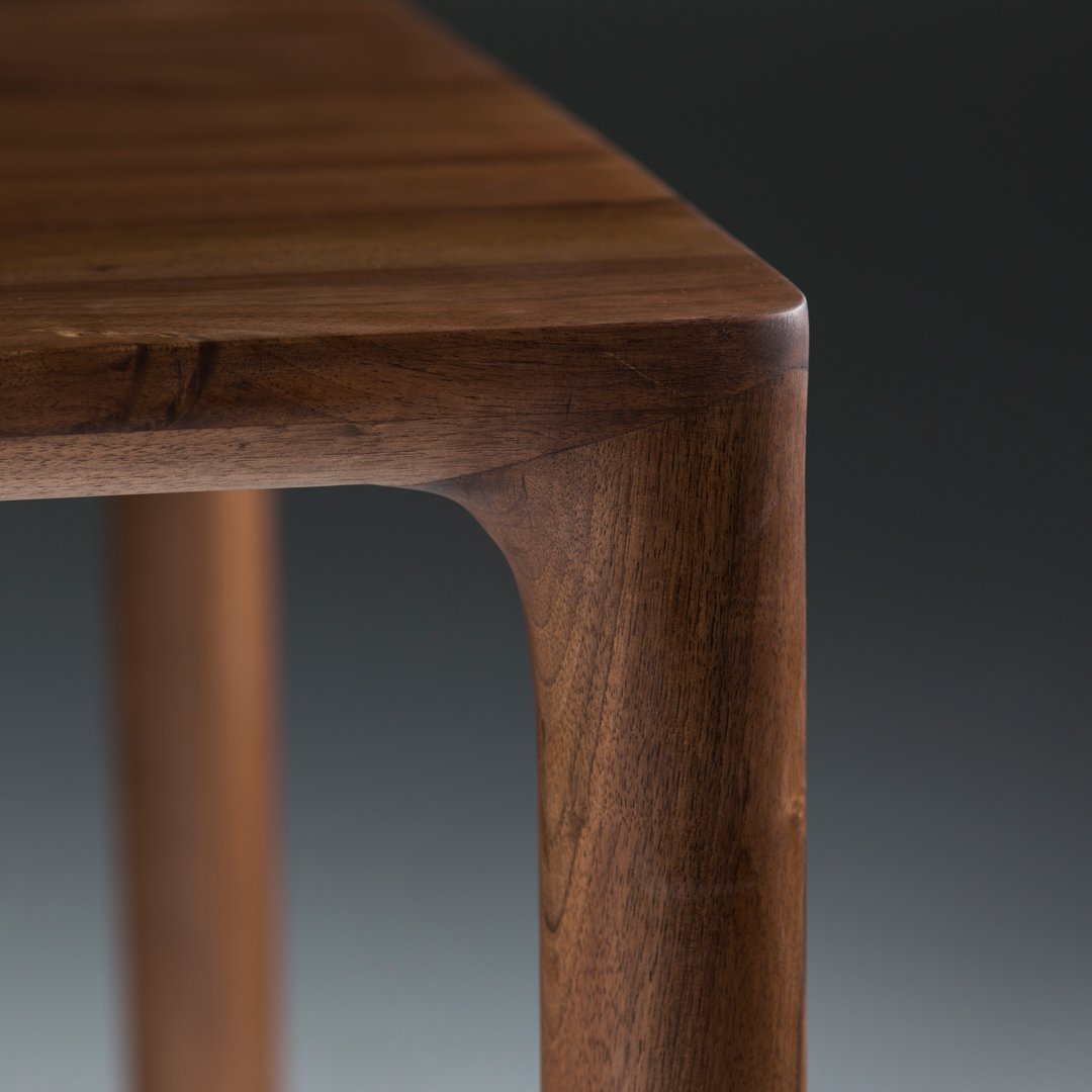 Close-up of rich brown wooden table leg with smooth curves and a natural grain pattern against a dark background.