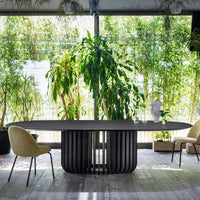 Black oval table with ribbed base, surrounded by green plants and light beige chairs, set against a bright, natural backdrop.