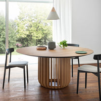 Light wood round dining table with natural grain pattern, surrounded by black and gray chairs in a bright space.