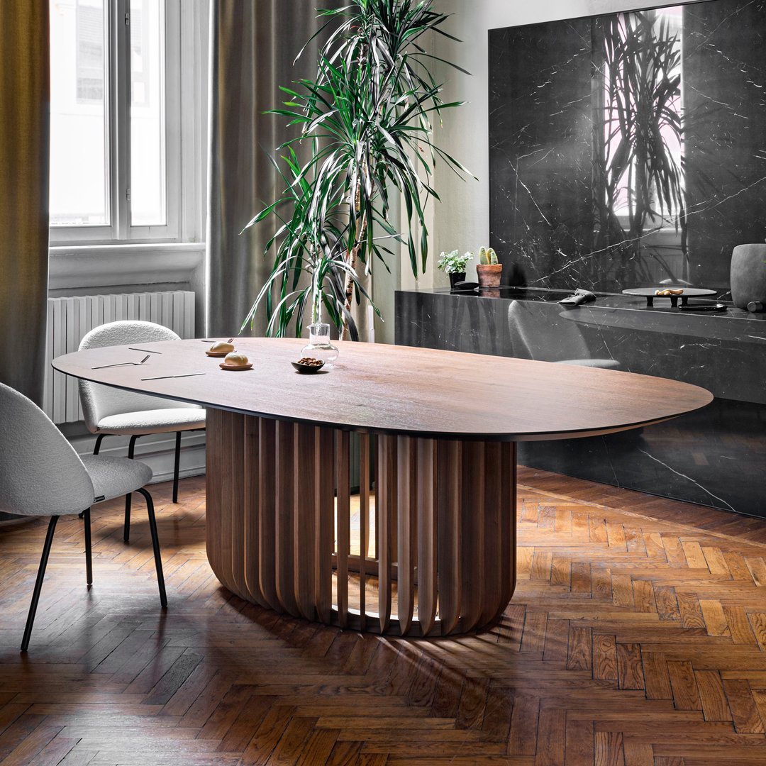 Elegant oval wooden table with a warm walnut finish, featuring a unique slatted base and rich herringbone flooring.