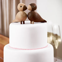 Wooden bird cake topper in natural brown shades, sitting atop a smooth white cake with a pink ribbon accent.